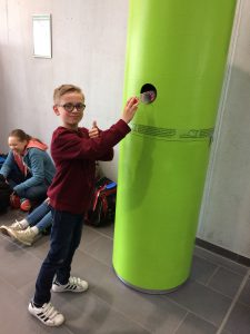 Ein Junge mit Brille steht neben einem großen, grünen Säulenobjekt und steckt seine Hand in ein Loch in der Säule. Er zeigt Daumen hoch und lächelt. Im Hintergrund sind weitere Kinder zu sehen, die sich unterhalten. Der Boden ist mit grauen Fliesen ausgelegt.