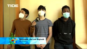 Drei junge Männer stehen nebeneinander vor einer Wand. Alle tragen Mund-Nasen-Masken. Der erste Mann auf der linken Seite trägt ein braunes Oberteil, der zweite trägt ein gestreiftes T-Shirt und der dritte trägt einen schwarzen Kapuzenpullover. Unter dem Bild befindet sich ein blauer Balken mit dem Text: 