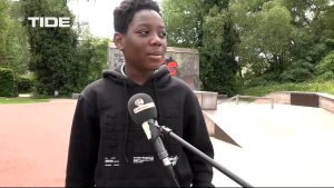 Junger Junge mit schwarzem Hoodie steht im Freien und spricht in ein Mikrofon. Im Hintergrund sind Bäume und eine Skateboard-Anlage zu sehen.