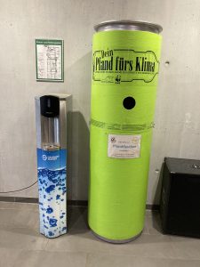 Wassertankstelle auf der linken Seite und ein hoher, grüner Behälter mit der Aufschrift „Dein Plan fürs Klima“ auf der rechten Seite. Der Hintergrund ist grau, und es gibt eine Wandkarte darüber.