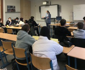 Eine Lehrerin steht vor einer Gruppe von Schülern in einem Klassenzimmer. Die Schüler sitzen an Tischen und hören aufmerksam zu. Einige Schüler tragen Masken. Im Hintergrund sind ein Whiteboard und ein Bildschirm zu sehen.