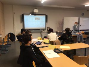 Eine Gruppe von Personen sitzt an Tischen in einem Klassenraum. Im Vordergrund sind mehrere Teilnehmerinnen und Teilnehmer sichtbar, die auf eine Präsentation an der Wand schauen. Eine Person sitzt auf einem Tisch und spricht, während ein Laptop und einige Unterlagen auf dem Tisch liegen. Der Raum ist hell und modern eingerichtet.