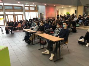 Eine Gruppe von Studierenden sitzt in einem modernen Raum mit großen Fenstern. Sie tragen Masken und sind auf Stühlen vor Tischen platziert. Im Hintergrund sind weitere Personen und die Raumgestaltung ist hell und einladend.
