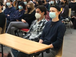 Zwei junge Männer sitzen an einem Tisch in einem Raum voller Menschen. Beide tragen Mund-Nasen-Schutzmasken. Der erste Mann hat lange, dunkle Haare und trägt ein kariertes Hemd, der zweite Mann hat kurze blonde Haare und trägt ein dunkelblaues T-Shirt. Im Hintergrund sind weitere Personen zu sehen, die ebenfalls an der Veranstaltung teilnehmen.