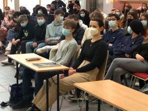 Eine Gruppe von Schülerinnen und Schülern sitzt in einem Raum. Einige tragen Mund-Nasen-Schutz. Im Vordergrund sind zwei Personen an einem Tisch zu sehen, eine Person sitzt auf einem Stuhl, während die andere auf einem weiteren Stuhl neben einem Tisch sitzt. Die Atmosphäre wirkt aufmerksam und konzentriert.