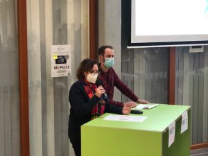 Zwei Personen stehen an einem grünen Tisch. Die Frau spricht ins Mikrofon und trägt eine Maske, während der Mann neben ihr steht und auf ein Dokument schaut. Im Hintergrund ist ein Bildschirm zu sehen.