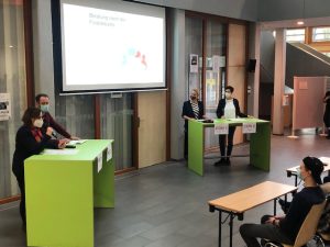 Zwei Tische in einem Raum, an denen Personen stehen. Eine Person spricht in ein Mikrofon, während andere Zuhörer sitzen. Im Hintergrund ist ein Bildschirm mit einer Präsentation zu sehen. Alle Anwesenden tragen Masken. Der Raum hat große Fenster und eine moderne Einrichtung.