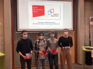 Vier Personen stehen in einem Raum und halten jeweils rote Objekte in den Händen. Sie tragen Masken. Im Hintergrund ist eine Präsentation zu sehen mit dem Text „WER MACHEN MIT!“ und „Handeln willkommen zum SchulEvent 2022 am Gymnasium Musterdorf“. Der Raum wirkt freundlich und modern.