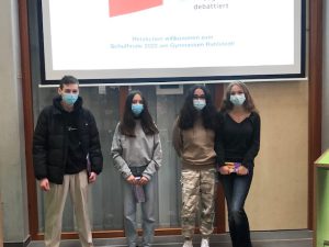 Vier Personen stehen nebeneinander in einem Raum, in dem eine Präsentation auf einer Leinwand im Hintergrund gezeigt wird. Alle tragen Mund-Nasen-Schutzmasken. Die Personen sind unterschiedlich gekleidet: Eine Person trägt eine schwarze Jacke, eine andere ist in grauem Sweatshirt und Hose, eine dritte trägt eine graue Jogginghose und die vierte Person hat einen schwarzen Pullover an. Die Leinwand zeigt einen roten Hintergrund mit Text, der teilweise unleserlich ist.