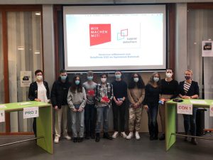 Gruppe von Jugendlichen mit Mund-Nasen-Schutz steht vor einer Präsentationsleinwand. Im Hintergrund sind zwei grüne Tische mit den Beschriftungen 