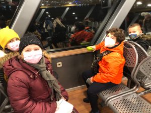Vier Kinder sitzen in einem Innenraum eines Fahrzeugs. Sie tragen Masken und warme Kleidung. Ein Mädchen mit einer gelben Mütze und ein Junge mit einer kurzen Frisur schauen in die Kamera, während ein anderes Kind in einem orangefarbenen Mantel am Fenster sitzt. Im Hintergrund sind weitere Passagiere und eine reflektierende Fensterfläche zu sehen.