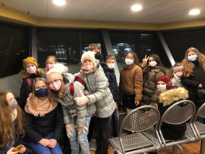 Gruppe von Kindern und Jugendlichen in einem Raum, die Masken tragen. Sie stehen und sitzen auf Stühlen, einige posieren fröhlich für das Foto. Der Raum hat große Fenster mit Blick auf die Außenbeleuchtung. Es ist Abend oder Nacht.