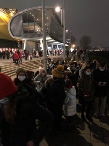 Eine Menschenmenge steht in einer Schlange vor einem modernen Gebäude bei Nacht. Viele Personen tragen Masken und warme Kleidung. Im Hintergrund sind Lichter und eine gelbe Plane sichtbar. Die Szene vermittelt eine Atmosphäre von Vorfreude und Aktivität.