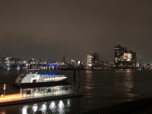 Nachtaufnahme eines Hafens mit einem beleuchteten Boot am Steg. Im Hintergrund sind beleuchtete Gebäude und ein schwach erhellter Himmel zu sehen. Die Wasseroberfläche spiegelt die Lichter wider.