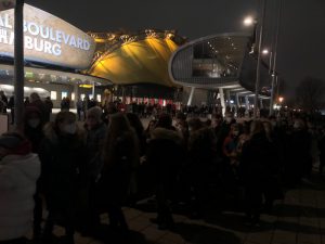 Eine große Menschenmenge steht an einem Veranstaltungsort in der Nacht. Im Hintergrund ist ein auffälliges Gebäude mit einem gelben, wellenförmigen Dach und der Beschriftung 
