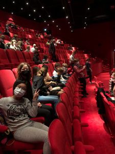 Eine Gruppe von Jugendlichen sitzt in einem roten Kinosaal. Einige tragen Masken. Die Reihen sind leer, bis auf die Gruppe, die sich entspannt unterhält und auf die bevorstehende Vorstellung wartet. Die Wände und Sitze des Saals sind in einem kräftigen Rot gehalten.