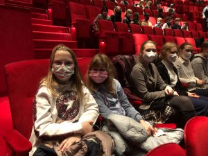 Zwei junge Mädchen sitzen in einem roten Kinosaal, beide tragen Masken. Im Hintergrund sind weitere Zuschauer auf roten Sitzen zu sehen. Die Atmosphäre ist entspannend und die Mädchen wirken fröhlich und aufgeregt.
