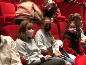 Drei Kinder sitzen in einem Kinosaal auf roten Sitzen. Sie tragen Gesichtsmasken und schauen in die Kamera. Im Hintergrund sind weitere Personen zu sehen, die sich bewegen.