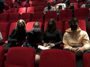 Vier Jugendliche sitzen in einem Kinosaal auf roten Sitzen. Alle tragen Mund-Nasen-Schutzmasken. Im Hintergrund sind weitere Personen zu sehen, die ebenfalls auf den Sitzen sitzen.