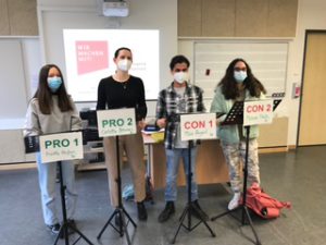 Vier Jugendliche stehen in einem Klassenraum vor einem Bildschirm. Sie tragen Mund-Nasen-Schutzmasken. Vor ihnen stehen Stative mit Schildern: zwei mit 