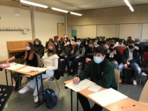 Eine Schülergruppe sitzt in einem Klassenraum. Die meisten tragen Masken. Die Tische sind mit Arbeitsmaterialien bedeckt, und im Hintergrund sind Stühle und ein Klavier zu sehen.