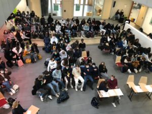 Eine große Gruppe von Menschen sitzt in einem Raum, der durch natürliche Beleuchtung erhellt wird. Die Anordnung zeigt zahlreiche Personen, die in verschiedenen Positionen sitzen, einige haben Rucksäcke dabei. Im Vordergrund sind einige Tische zu sehen. Der Raum hat eine offene, einladende Atmosphäre.
