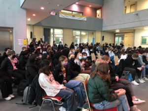 Eine große Gruppe von Schülern sitzt in einem Innenraum. Viele tragen Masken. Der Raum hat helle Wände und große Fenster. Einige Schüler sind auf Stühlen, während andere auf dem Boden sitzen. Im Hintergrund sind weitere Personen und eine Bannerabhängung zu sehen.