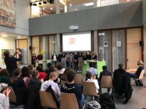 Eine Gruppe von Schülern steht auf einer Bühne vor einem Bildschirm, während Zuschauer im Publikum sitzen. Der Raum ist hell erleuchtet und hat eine moderne Architektur. Im Hintergrund sind weitere Schüler und Lehrer zu sehen, die aufmerksam der Präsentation folgen.