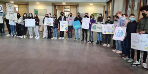 Gruppenschülerinnen und -schüler stehen in einem Raum und halten Plakate mit verschiedenen Zeichnungen und Slogans. Alle tragen Masken. Der Raum hat helle Wände und ist gut beleuchtet.