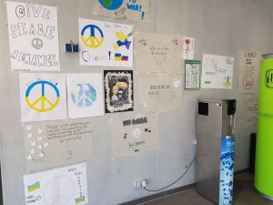 Wand mit verschiedenen Plakaten und Zeichnungen, die Frieden, Hoffnung und Unterstützung für die Ukraine thematisieren. Auf den Plakaten sind Frieden-Symbole, Texte wie 