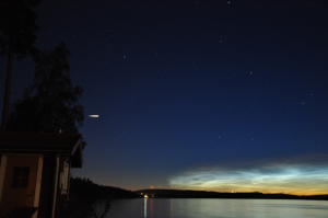 Nachtaufnahme eines ruhigen Sees, umgeben von Bäumen und Hügeln. Der Himmel ist tiefblau mit zahlreichen sichtbaren Sternen. Am Horizont ist ein sanfter Lichtschein zu sehen, der auf den Sonnenuntergang hinweist. Links im Bild steht ein kleines Gebäude, während am rechten Rand des Bildes eine helle Wolke zu erkennen ist.