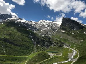 Eine malerische Berglandschaft mit grünen Hügeln, schneebedeckten Gipfeln und einem blauen Himmel mit einigen Wolken. Im Vordergrund schlängelt sich ein Weg durch die Wiesen, während Wasserfälle die Hänge hinunterfließen.