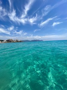 Klares, turquoise Wasser mit sanften Wellen, unter einem blauen Himmel mit wispy Wolken. Am Horizont sind sanfte Berge und eine Küstenlinie mit Gebäuden sichtbar.