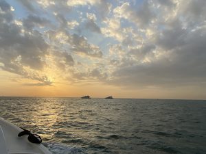 Ein Sonnenuntergang über dem Meer, mit sanften Wellen und einem bewölkten Himmel. Im Hintergrund sind zwei kleine Boote sichtbar. Die Farben des Himmels variieren von warmen Gelbtönen bis zu kühleren Grautönen. Am unteren Bildrand ist ein Teil des Bootes zu sehen.