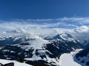 Panoramablick auf schneebedeckte Berge unter einem klaren blauen Himmel mit einigen Wolken. Im Vordergrund erstreckt sich ein schneebedeckter Hang, während ein gefrorener See im Tal sichtbar ist. Die Landschaft vermittelt ein Gefühl von Weite und natürlicher Schönheit.