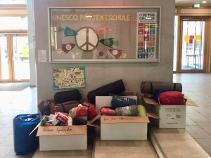 Im Eingangsbereich einer Schule steht ein Tisch mit mehreren Kartons und Taschen. Die Kartons sind mit verschiedenen gespendeten Materialien gefüllt. Hinter den Kartons hängt ein Plakat mit dem Schriftzug 