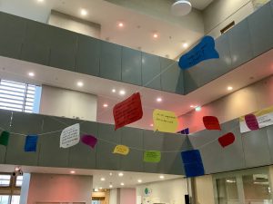 Innerhalb eines modernen Innenraums mit hellen Wänden und großen Fenstern hängen farbige Sprechblasen aus Papier von der Decke. Die Sprechblasen sind in verschiedenen Farben gestaltet und enthalten Texte, die möglicherweise Zitate oder Botschaften darstellen. Der Raum hat eine offene Architektur mit mehreren Ebenen und wird von natürlichem Licht durchflutet.