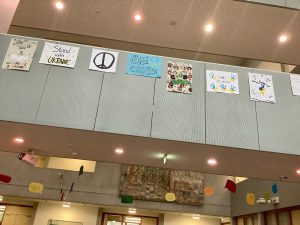 In einem Innenraum sind mehrere bunte Plakate an einer Wand aufgehängt. Die Plakate sind unterschiedlich gestaltet und enthalten Texte und Zeichnungen, die Frieden und Unterstützung für die Ukraine thematisieren. Der Raum hat eine helle Beleuchtung, und es sind farbige Dekorationen an der Decke sichtbar.