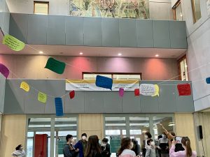 Eine Gruppe von Menschen steht in einem modernen, hellen Raum mit großen Fenstern. An den Wänden sind bunte Sprechblasen und ein Banner angebracht. Im Hintergrund ist ein großes Wandgemälde zu sehen. Die Personen scheinen an einer Veranstaltung oder Aktivität teilzunehmen.