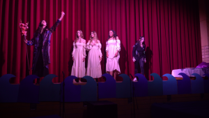 Eine Theateraufführung mit vier Darstellerinnen auf der Bühne. Eine Schauspielerin in schwarzer Kleidung hält einen Blumenstrauß und gestikuliert nach oben, während die anderen drei Darstellerinnen in weißen Kleidern stehen. Im Hintergrund ist ein roter Vorhang sichtbar. Bunte Wellen aus Pappe sind am Bühnenrand platziert.