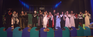 Eine Gruppe von Schauspielern steht auf einer Bühne und applaudiert. Sie tragen verschiedene Kostüme, darunter Piraten- und historische Gewänder. Im Hintergrund sind Bühnenelemente wie Pflanzen und Wellen zu sehen. Die Beleuchtung ist gedämpft und es herrscht eine festliche Atmosphäre.