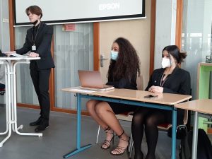 Eine Person steht an einem Tisch und präsentiert, während zwei weitere Personen an einem Tisch sitzen. Alle tragen Mund-Nasen-Schutz. Die Präsentation findet in einem Raum mit einem Bildschirm im Hintergrund statt.