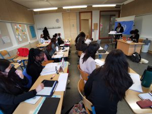 Eine Gruppe von Studierenden sitzt in einem Klassenraum an Tischen und arbeitet an Laptops oder Notizen. Im Vordergrund sind mehrere Personen zu sehen, die auf ihre Materialien konzentriert sind. Im Hintergrund steht eine Person an einem Pult und präsentiert vor einer Wand mit einem Banner. Der Raum ist hell und freundlich gestaltet, mit verschiedenen Materialien an den Wänden.
