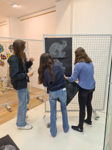 Drei Jugendliche stehen vor einer Stellwand, auf der ein Kunstwerk abgebildet ist. Sie sind mit Stiften oder Farben beschäftigt und scheinen an einem kreativen Projekt zu arbeiten. Die Umgebung wirkt hell und freundlich, mit Holzfußboden und einer weiteren Stellwand im Hintergrund, die mit bunten Kunstwerken dekoriert ist.