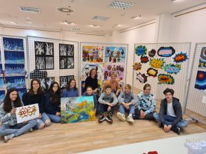 Gruppe von Jugendlichen und Erwachsenen sitzt auf dem Boden in einem Kunstausstellungsraum. Im Hintergrund sind verschiedene Kunstwerke und Plakate an Wänden ausgestellt. Einige Personen halten Bilder in den Händen. Der Raum hat helle Wände und einen Holzfußboden.