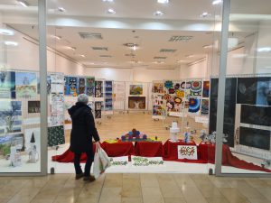 Eine Person mit einem Einkaufsbeutel steht vor einer Glasfront, die den Innenraum einer Galerie zeigt. Im Raum sind zahlreiche Kunstwerke an den Wänden ausgestellt, darunter Gemälde und andere kreative Arbeiten. Der Boden ist mit roten Tüchern dekoriert, und einige Objekte sind aufgestellt. Die Beleuchtung ist hell und freundlich.