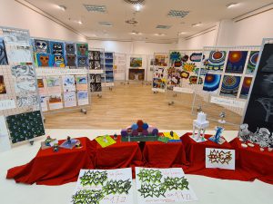 Ein Raum mit verschiedenen Kunstwerken und Ausstellungsstücken. An den Wänden hängen zahlreiche farbige Poster und Bilder. Im Vordergrund stehen Tische mit roten Tischdecken, auf denen diverse Objekte und bunte Materialien ausgestellt sind. Der Boden ist aus Holz und der Raum ist gut beleuchtet.