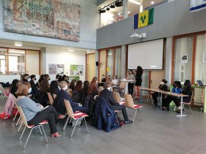 Eine Gruppe von Menschen sitzt in einem Raum und hört einer Präsentation zu. Im Vordergrund steht eine Frau, die spricht, während im Hintergrund eine Leinwand zu sehen ist. An den Wänden hängen verschiedene Flaggen, und der Raum hat eine moderne, offene Gestaltung.