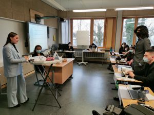 Eine Unterrichtssituation in einem Klassenraum. Eine Person steht vorne und präsentiert, während mehrere Schüler an Tischen sitzen und auf Laptops arbeiten. Alle Personen tragen Masken. Im Hintergrund sind große Fenster, durch die Tageslicht fällt.
