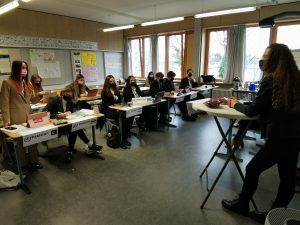 Eine Gruppe von Schülern sitzt an Tischen in einem Klassenzimmer. Vor ihnen steht eine Lehrperson, die einen Unterrichtsstoff präsentiert. Alle Anwesenden tragen Masken. Das Klassenzimmer ist hell erleuchtet, mit Fenstern, die Tageslicht hereinlassen, und einer Tafel im Hintergrund.